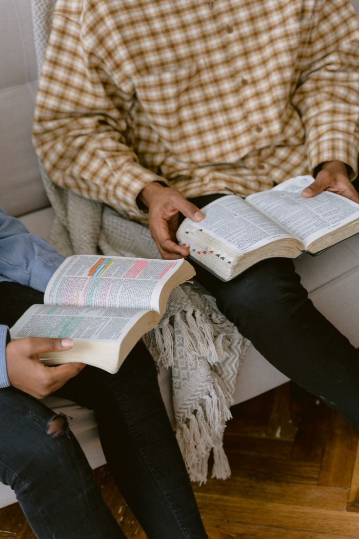 Two individuals sitting on a sofa reading and discussing the Bible indoors.