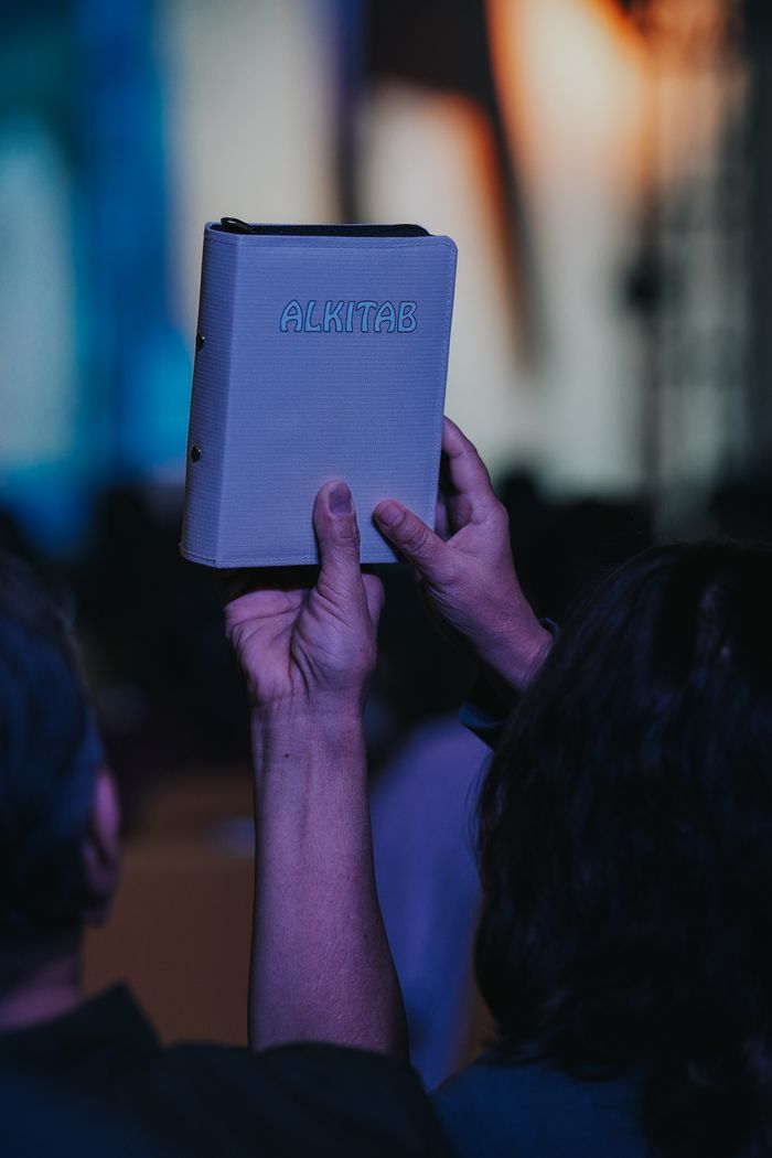 about-us-01 A person holds up an Alkitab Bible during a worship service, symbolizing faith and devotion.